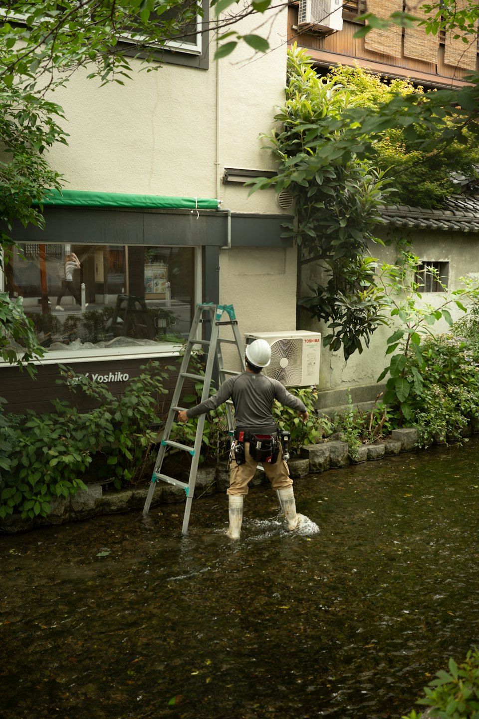 Technician in waders holds a ladder while repairing an air conditioner.